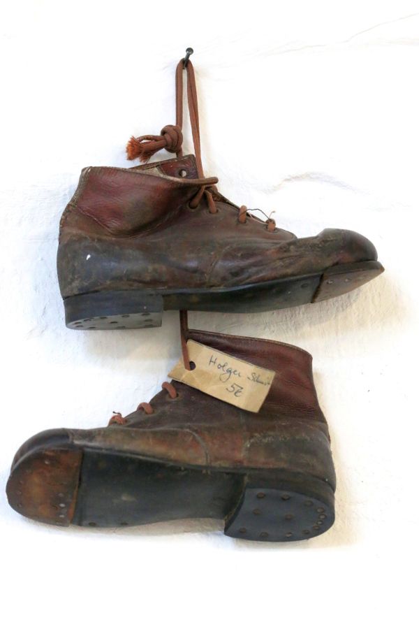 Das Bild zeigt ein Paar altmodische, abgenutzte Schuhe mit abgerundeter Vorderkappe, die deutliche Gebrauchsspuren und eine robuste, genagelte Sohle aufweisen; an einem Schuh hängt ein Etikett mit der Aufschrift „Holger Schuh SE