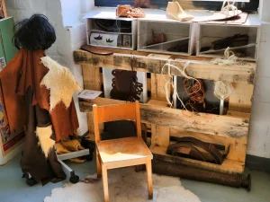 Schuhmacher-Arbeitsplatz mit rustikaler Holzwerkbank, hängenden Schuhformen und Lederteilen, weißen Regalen mit Werkzeugen im Hintergrund, einer Schaufensterpuppe mit braunem Ledergewand links und einem einfachen Holzstuhl - traditioneller Handwerksarbeitsbereich im Museum mit natürlichem Charme.