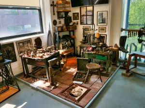 Vollständige Schuhmacherwerkstatt-Szene im Museum mit mehreren Arbeitstischen, historischen Werkzeugen, Schuhleisten, Nähmaschinen, gerahmten Bildern an der Wand, einem Vogelkäfig und einem alten Foto auf dem Boden - authentische Rekonstruktion einer traditionellen Schuhmacherei mit Tageslicht durch die Fenster.