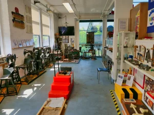 Werkstatt-Ausstellungsraum im Schuhmuseum mit historischen Schuhmachermaschinen, Nähmaschinen, roten Stufen-Exponaten und Vitrinen mit Werkzeugen und Materialien - ein interaktiver Bereich zur Demonstration des traditionellen Schuhmacherhandwerks.