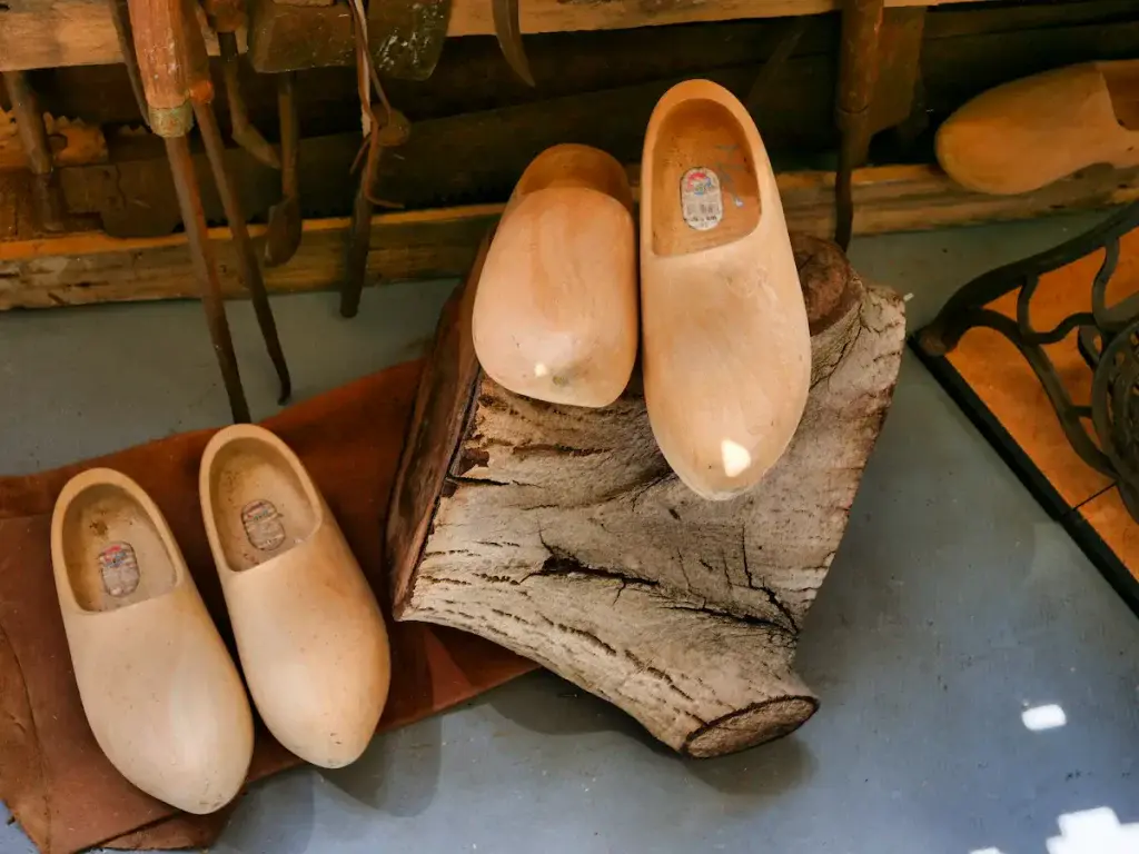 Holzclogs in verschiedenen Größen auf Birkenstämmen arrangiert - traditionelle Holzschuhe in einem Museums-Werkstatt-Setting mit historischen Werkzeugen im Hintergrund.