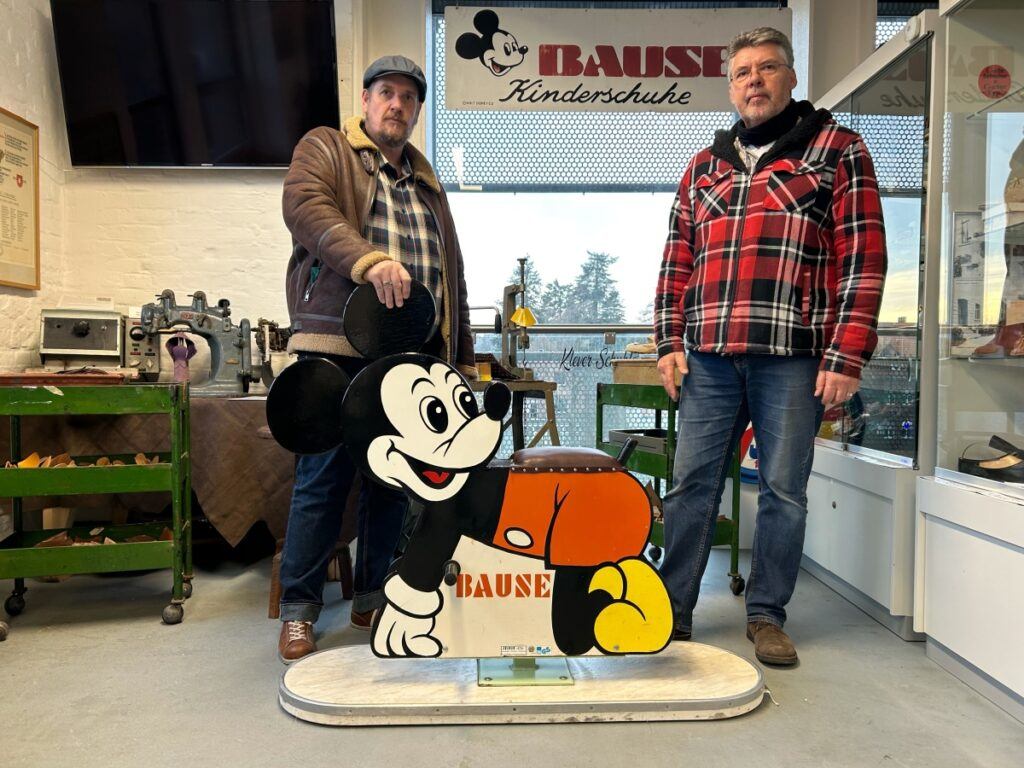 Mickey-Mouse-Pappfigur von "BAUSE Kinderschuhe" im Schuhmuseum vor historischen Exponaten und dem Markenschild.