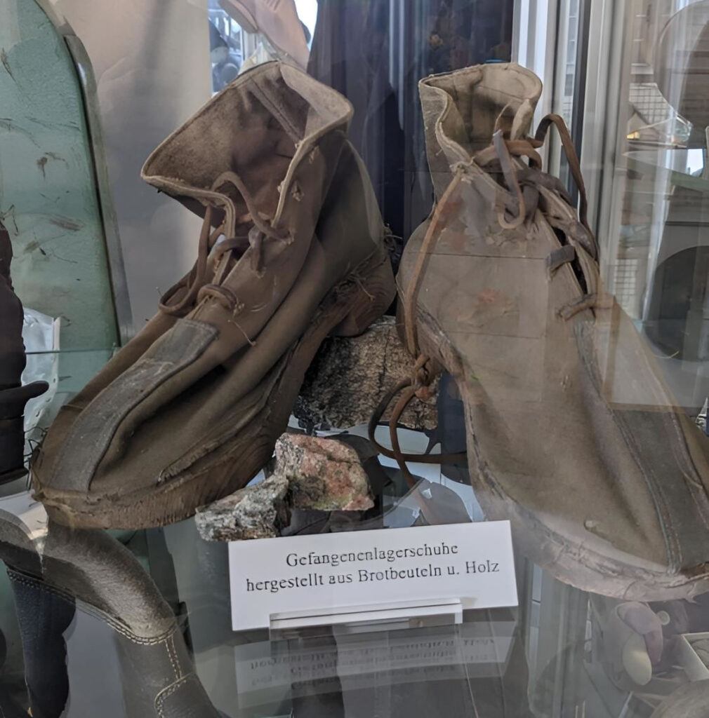 Historische Gefangenenlagerscuhe aus Brotbeuteln und Holz in einer Museumsvitrine - notdürftig hergestelltes Schuhwerk aus der Kriegs- oder Nachkriegszeit.