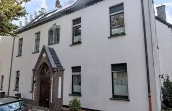 Weißes zweigeschossiges Wohnhaus mit neogotischem Eingangsportal und dunkel gerahmten Fenstern.