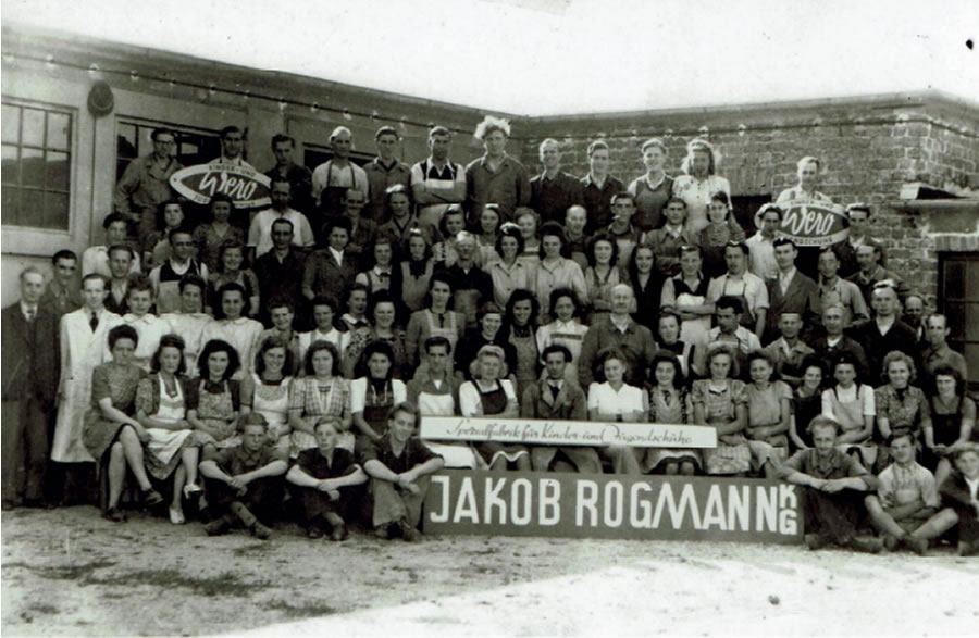 Historisches Belegschaftsfoto der Schuhfabrik Jakob Rogmann mit zahlreichen Mitarbeitern in Arbeitskleidung vor dem Fabrikgebäude.